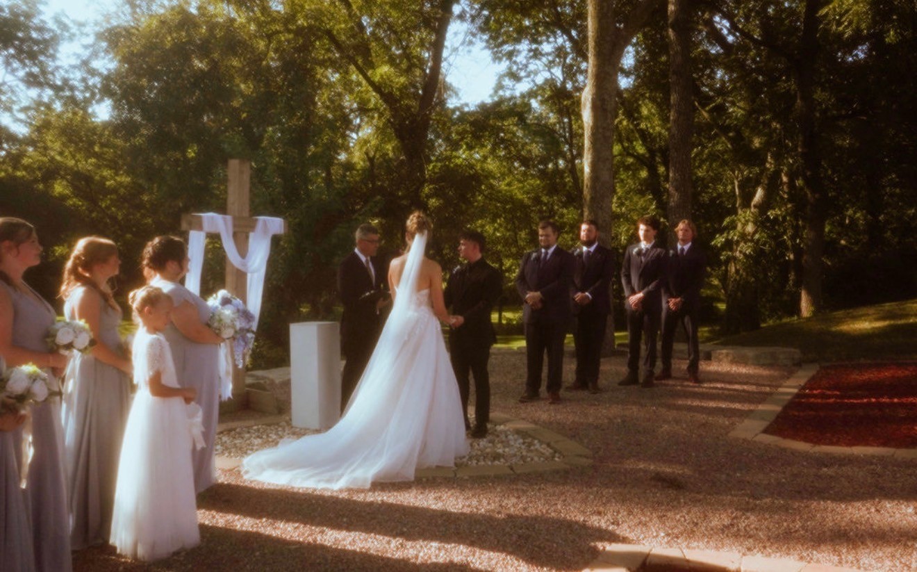 The bride stands with the groom at an outdoor ceremony under the trees, a wooden cross behind them