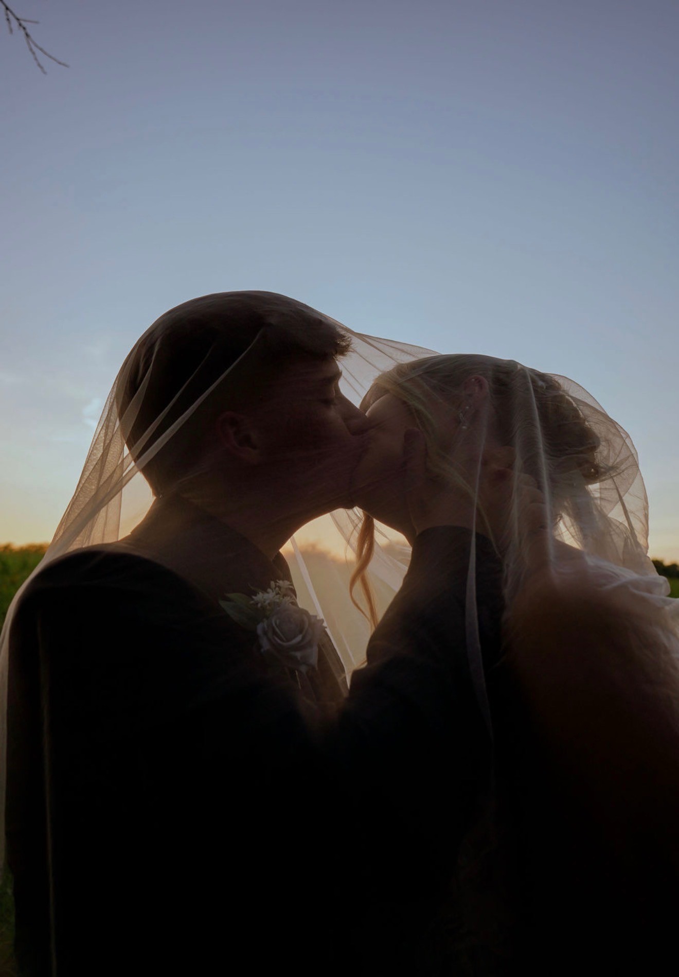 The couple kisses under a sheer veil, silhouetted against the evening sky
